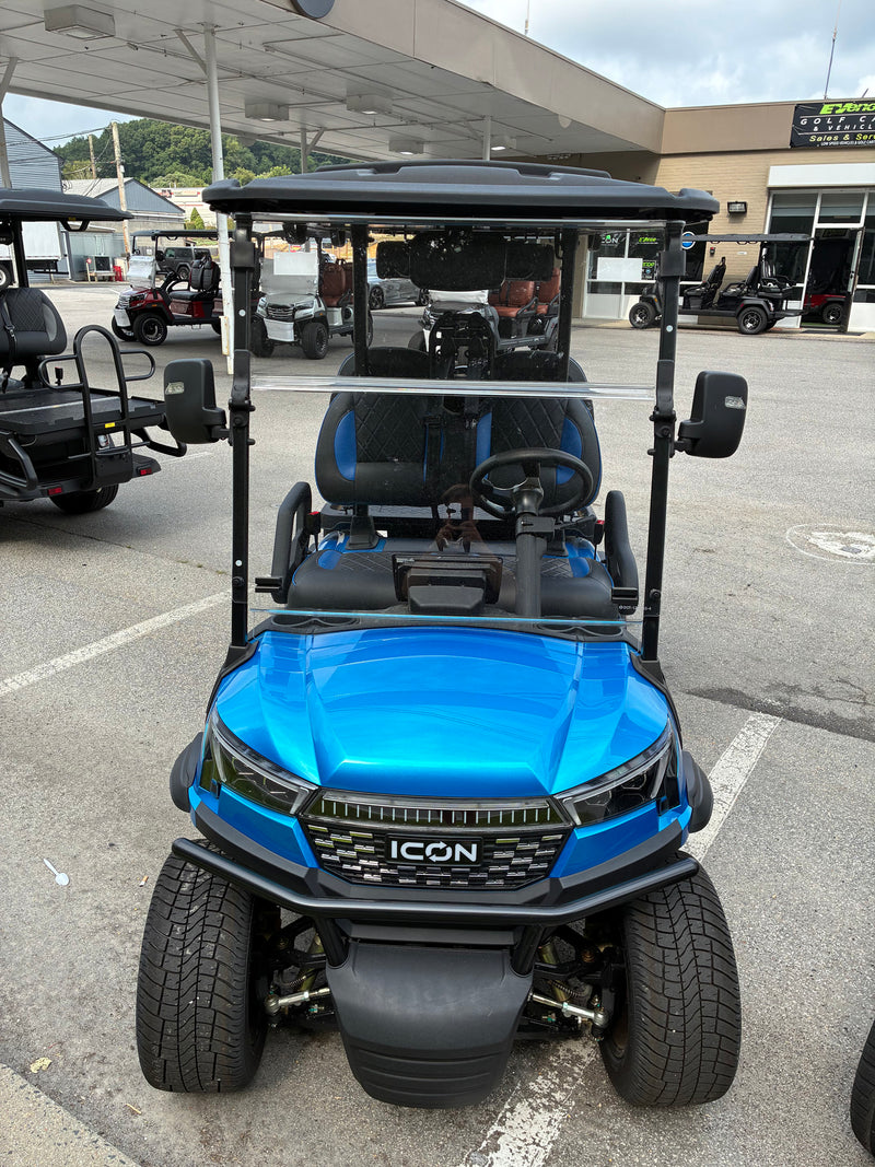 Load image into Gallery viewer, A bright blue and black ICON golf cart parked in an outdoor dealership lot. The cart features a black canopy, black seats with blue side bolsters, a black steering wheel, and a black front grille with the 'ICON' logo.