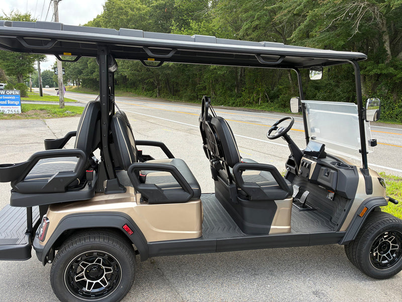 Load image into Gallery viewer, A four-seat golf cart parked outdoors on a paved road with trees and a road sign in the background. The cart has a beige body, black seats, a black roof, a clear windshield, and black wheels.