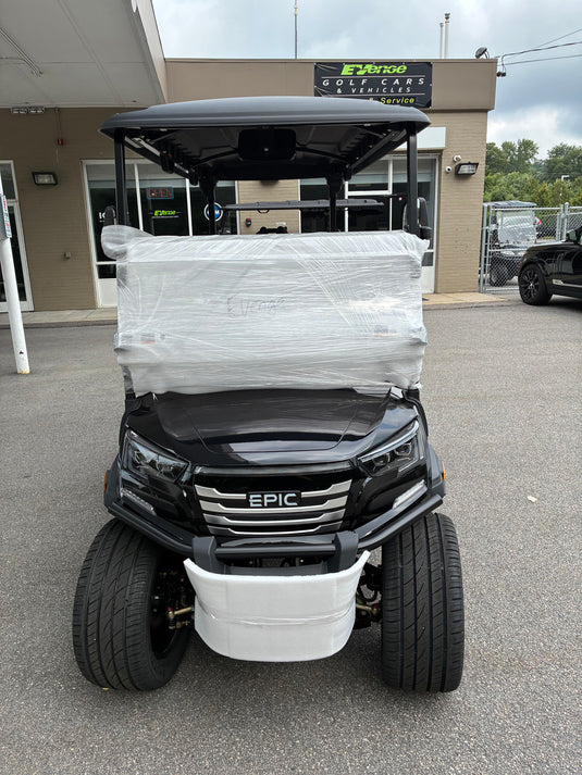 A black EPIC brand golf cart parked outdoors in front of a building, wrapped in clear plastic on the front cargo bed. The cart has a black canopy, black tires, and a white front bumper.
