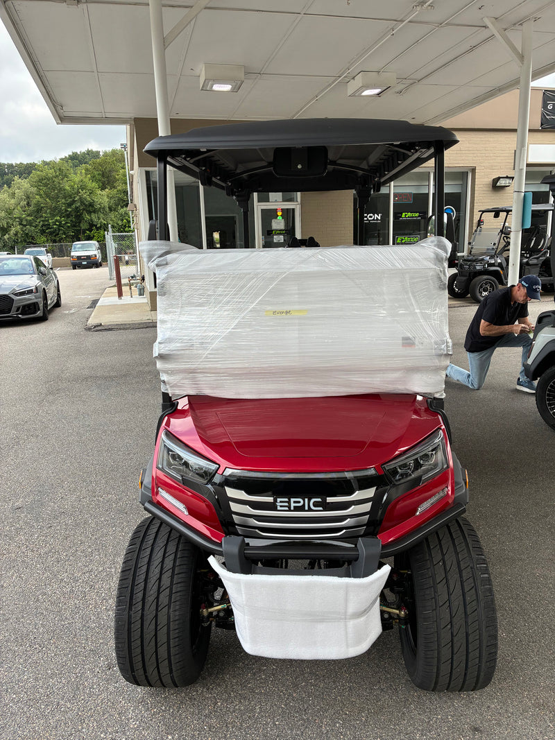 Load image into Gallery viewer, A red EPIC electric golf cart under a black canopy, wrapped in protective plastic on the front, photographed outdoors at a dealership with other carts and a building in the background.
