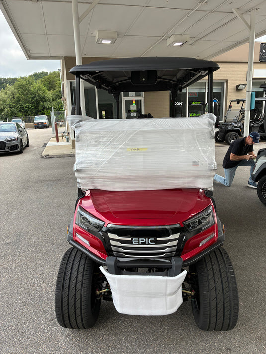 A red EPIC electric golf cart under a black canopy, wrapped in protective plastic on the front, photographed outdoors at a dealership with other carts and a building in the background.