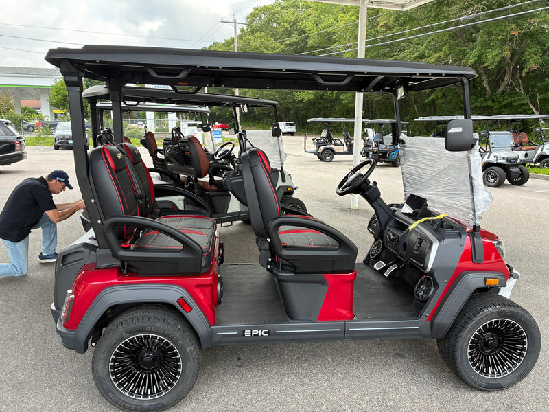 Load image into Gallery viewer, A red and black electric golf cart with 'EPIC' branding on the side, parked outdoors on a paved lot with other golf carts and a person kneeling beside it.
