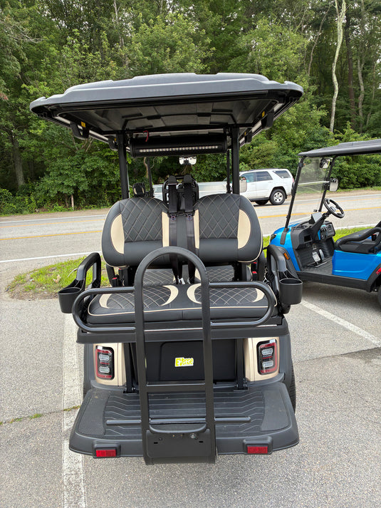 A rear view of a black and beige luxury golf cart with a hardtop canopy, parked on an asphalt lot next to another blue and black golf cart. Trees and a road are visible in the background.