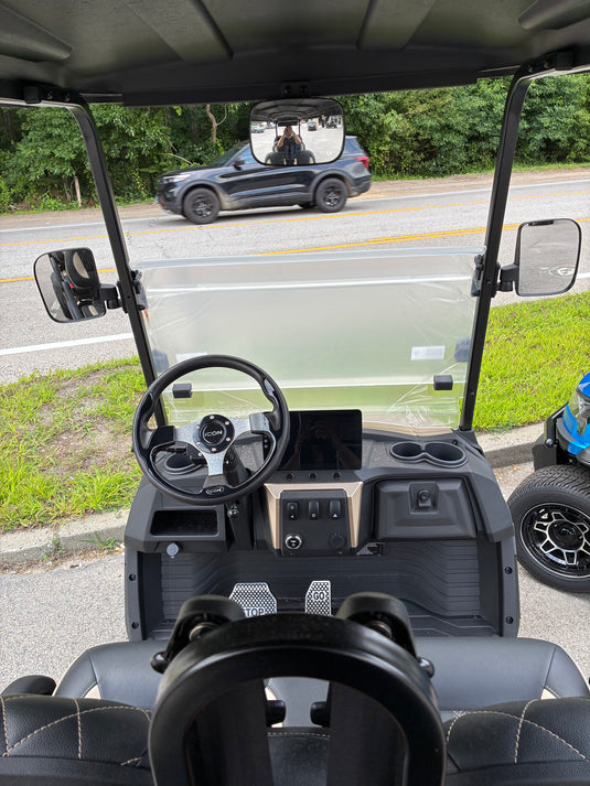 Black electric golf cart with a clear windshield, showing the interior dashboard, steering wheel, and side mirrors, photographed outdoors on a road with grass and trees in the background.