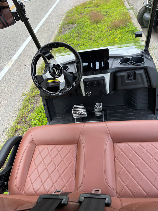 A luxury electric golf cart with a prominent quilted pink-brown leather front seat, black steering wheel, and dashboard, photographed from a high angle on a paved road next to grass.
