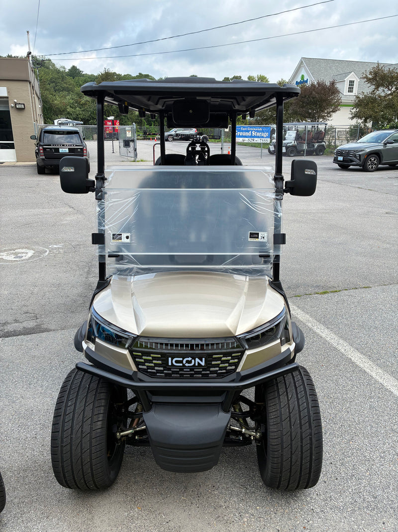 Load image into Gallery viewer, A gold and black ICON golf cart parked in an outdoor lot, with a clear protective plastic cover on the windshield.

