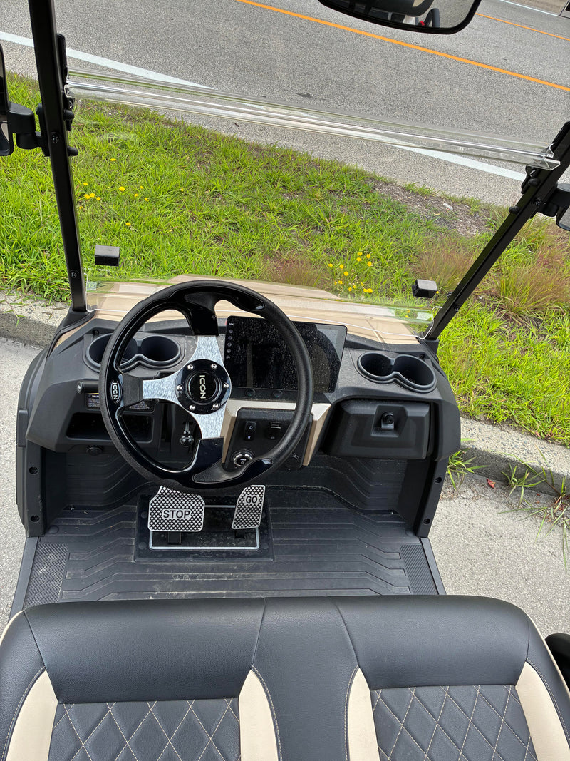 Load image into Gallery viewer, Front-facing view inside an electric golf cart, showing the black steering wheel, dashboard, foot pedals, and black-and-cream leather seats, parked outdoors on pavement beside grass and a road.
