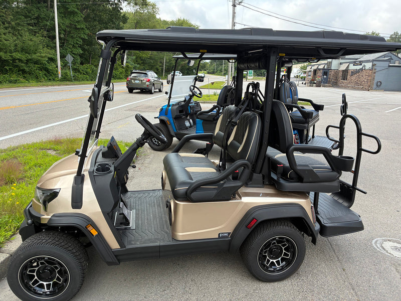 Load image into Gallery viewer, A beige and black electric golf cart with a four-passenger roof canopy, parked on an outdoor paved area next to a road with trees in the background.

