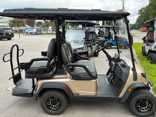 A luxury electric golf cart in champagne beige and black, parked outdoors in a dealership lot with other carts visible in the background.