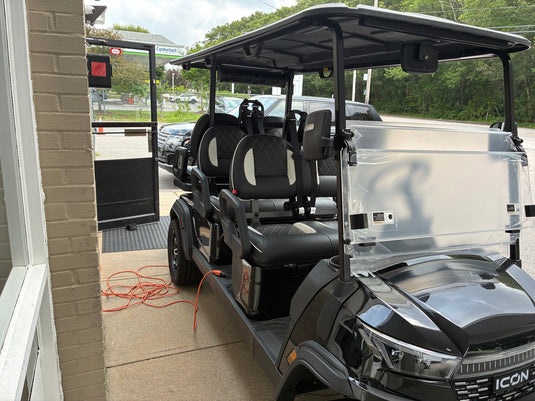 A black and gray six-passenger electric golf cart with a canopy, parked on a concrete driveway next to a brick building. The cart has a protective clear plastic sheet covering the rear side panel, and an orange extension cord is coiled on the ground nearby.
