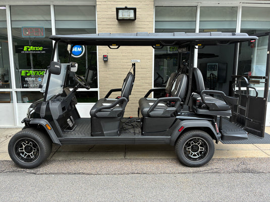 A black four-passenger electric golf cart parked outdoors in front of a building with large windows and glass doors. The cart has a dark canopy roof, black seats, and black wheels with silver rims.