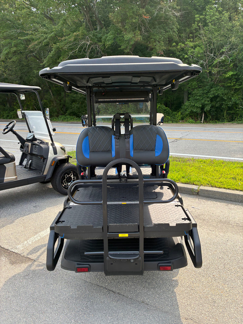 Load image into Gallery viewer, A black electric golf cart with two blue-accented seats, a roof canopy, and a rear cargo bed, photographed outdoors on a paved area with trees and another cart visible at the left edge.
