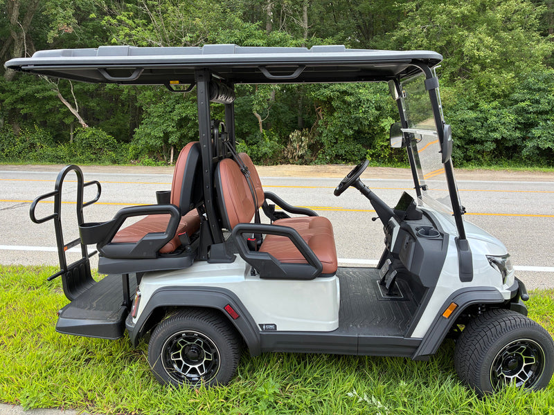 Load image into Gallery viewer, A white and black electric golf cart with brown leather seats, parked on a grassy area next to a road with trees in the background.
