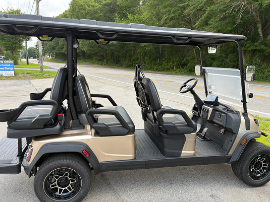 A four-seat golf cart parked outdoors on a paved road with trees and a road sign in the background. The cart has a beige body, black seats, a black roof, a clear windshield, and black wheels.